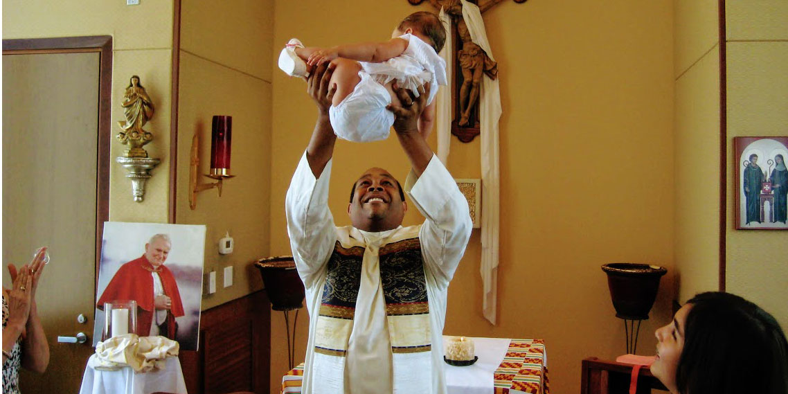 A photo of Drs. Lee Hobbs, Allyson Marino, their daughter Lila, and Fr. Stephan Brown in the chapel at University Campus when she was christened as a baby in 2011