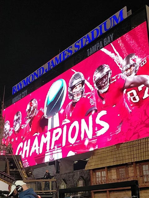 A photo showing the Jumbotron inside Raymond James Stadium in Tampa, FL on which it shows a few Tampa Bay Buccaneers players with the word 'Champs' and the pirate ship next to the screen.