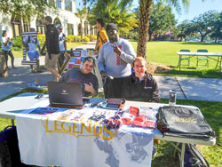 A photo of three students who have set up a table outside the SAB on University Campus promoting the eSports Club with giveaway items on the table and one of the students is playing a game on a laptop