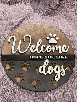 A photo of a circular wooden ornament that says ‘Welcome, Hope you Like Dogs’ with small paw cutouts; ; this was designed by Saint Leo University business administration degree student Brianna Adeeb for her Etsy store