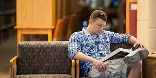 A photo of a young male college student reading a book in the library to study for a class for the bad study habits and how to break them blog article