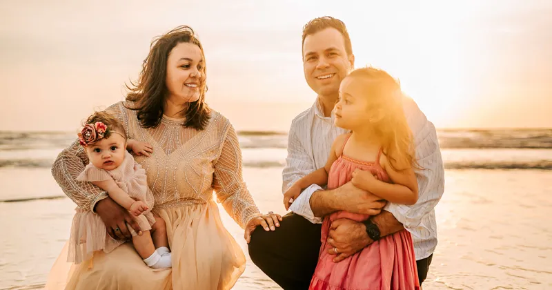 A photo of the Loubriel family smiling at the beach; Joanne and Javier are kneeling, and their daughters, Jade and Jolie, are standing; the water and sand are visible in the background