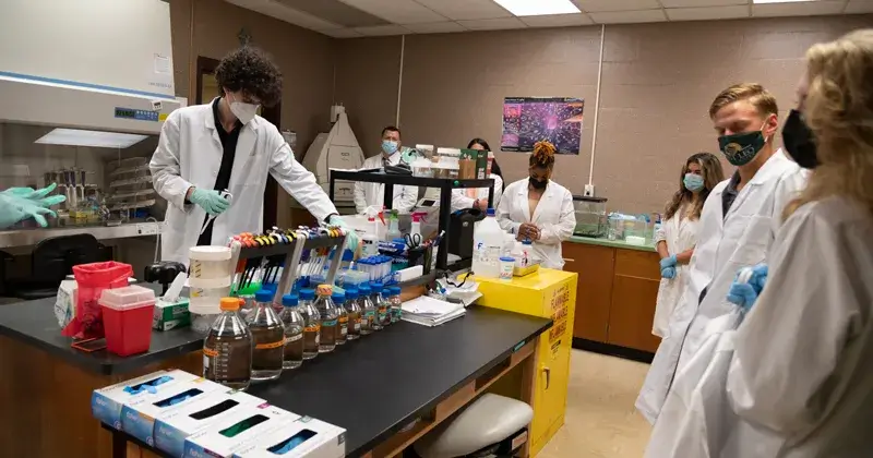 A photo of six Saint Leo University students in a biology lab; they are each wearing a lab coat and face mask; there are bottles and gloves on the counter in front of them; this is for the best biology apps for students blog article for those enrolled in a biology degree program