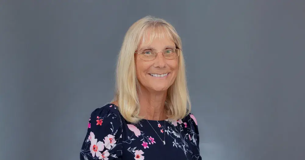 A head shot of Dr. Melinda “Lin” Carver, a full professor of graduate studies in education at Saint Leo University, smiling and wearing a necklace with a pendant hanging down and thin-rimmed eyeglasses; Carver is a Caucasian woman with long blond hair wearing a blue dress with bright-colored flowers on it; she teaches in the online graduate education program tracks for Saint Leo