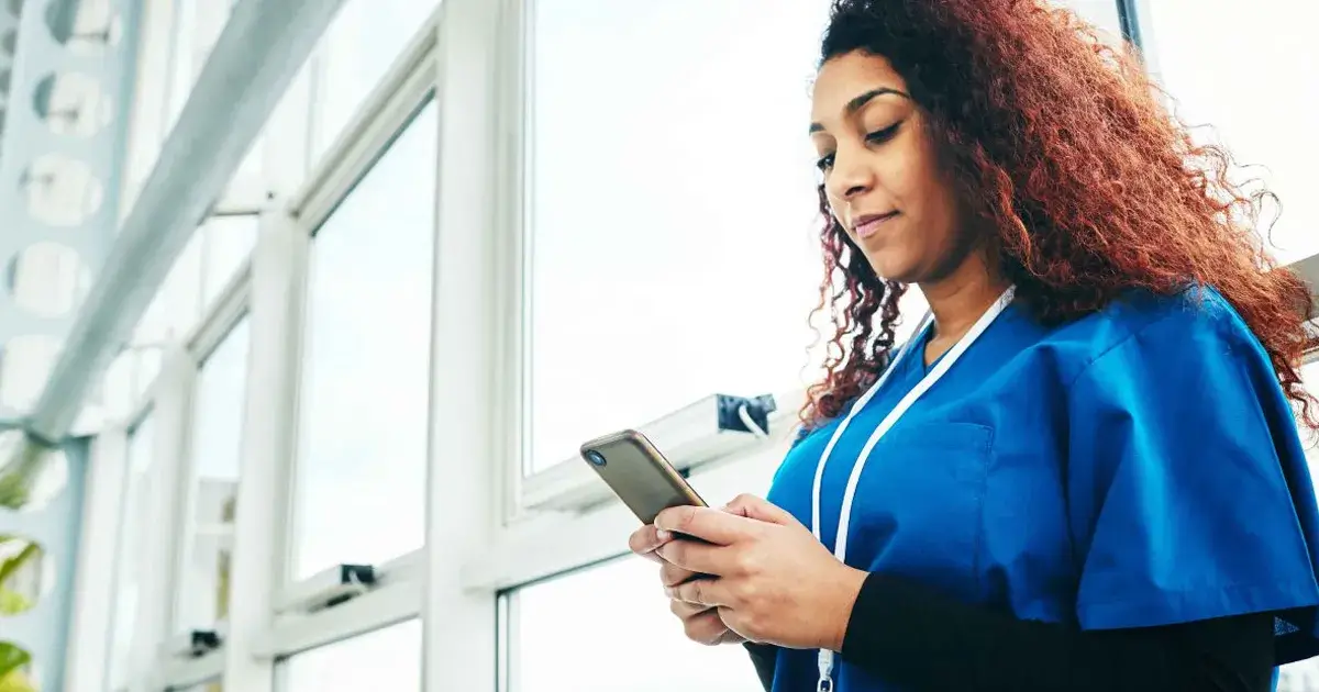 A photo of a female African-American nurse with curly hair wearing blue scrubs with a black undershirt standing in front of some windows and using a smartphone for the blog article on the best mobile apps for nursing students