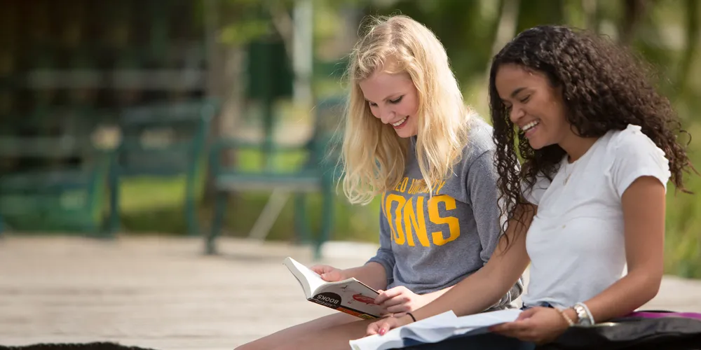 A photo of two female Saint Leo University students (one with blond hair and the other with brown hair) studying together outside; they are sitting on a bench with trees in the background; this is for the blog article on the benefits of having a study buddy in college