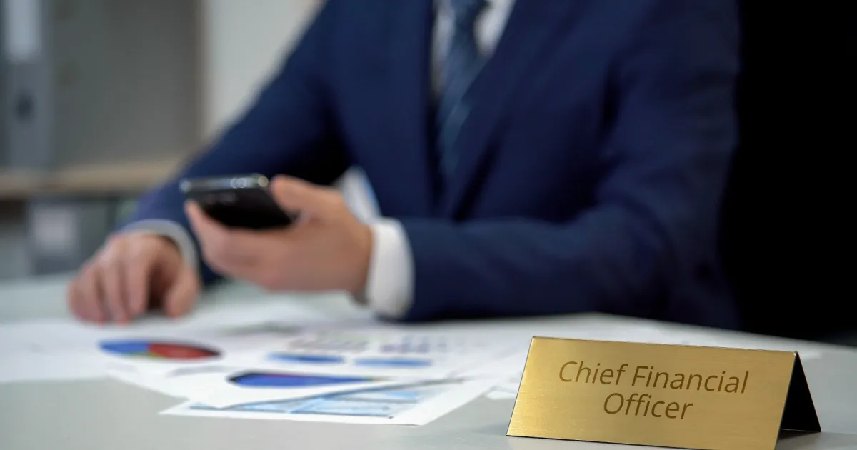 A photo of a Caucasian man sitting at a desk dressed in a business suit looking at his smartphone; in front of him is a nameplate that says 'Chief Financial Officer' and some graphs, charts, and other documents are lying on the desk; this is for the blog article on 'What does a CFO do?' 'How much does a CFO make?' and what the typical CFO job responsibilities actually entail