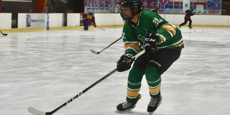 An action shot of Jack Prieto, a Saint Leo cybersecurity major in his senior year, skating from left to right on the ice with his stick in a club hockey game with the Saint Leo Lions' club hockey team which he helped establish in 2022; he is wearing a green Saint Leo uniform with yellow stripes as well as a black helmet