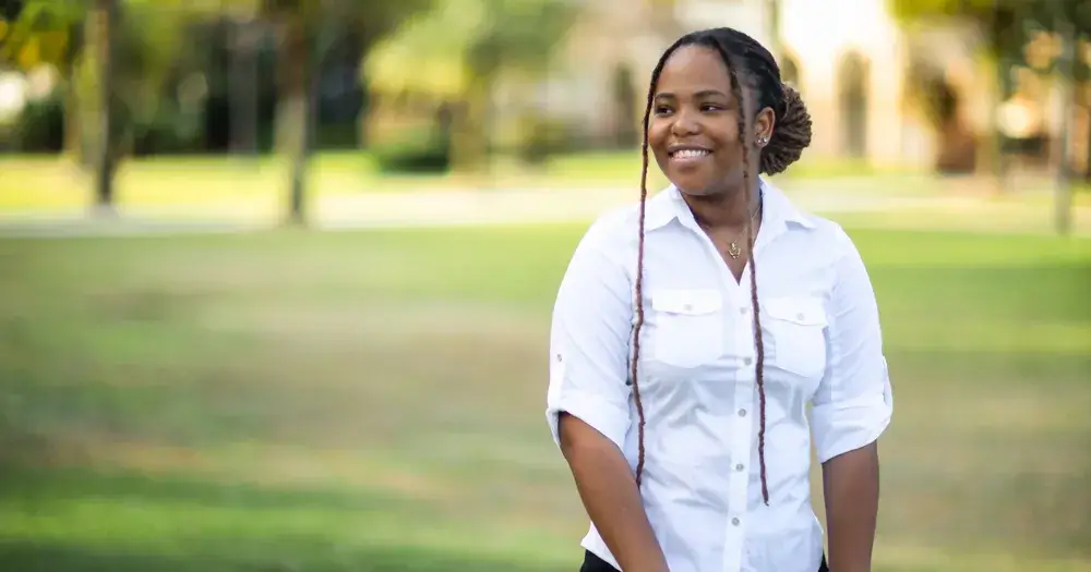 A photo of Tayhana Taylor, a Saint Leo University accounting major, standing on campus; she is looking to her right with a nice smile and is wearing black pants and a white shirt; University Campus is visible in the background