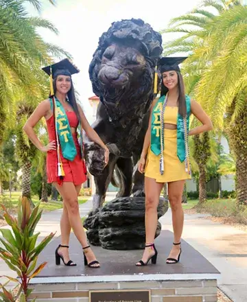 A photo of Jenna and Jessica Skrelunas, identical twins and Saint Leo alumni, smiling for a photo outside on University Campus next to the lion statue