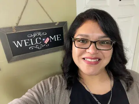 A head shot of Emily Gilbert, an alumna of the bachelor’s in psychology and Master of Social Work degree programs at Saint Leo University, wearing glasses and smiling; she is standing next to a sign on the wall that says ‘Welcome’ with the ‘o’ in the shape of a heart and an American flag inside of it