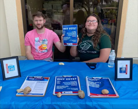 A photo of Megan Rowe, a current undergraduate social work degree student at Saint Leo University, with a male student at a table on University Campus handing out flyers for the ‘Why Vote?’ campaign to encourage students to vote