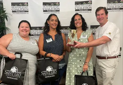 A photo of Mary Martinez-Drovie, Maria Dacosta, and Julia Sharp receiving their award and swag bags from Dr. Mark Gesner, vice president of Community Engagement and Communications at Saint Leo University, for placing first in the ‘A Solution That Matters’ category of the ‘What Matters on Mane Street’ social impact competition held by Saint Leo in 2022