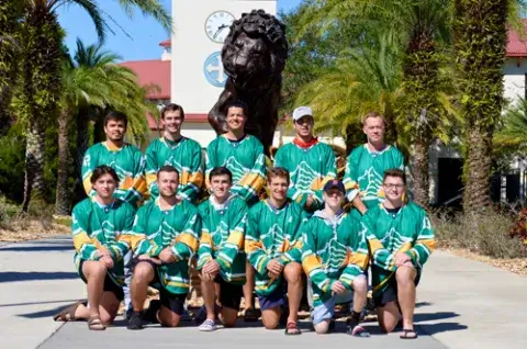 A team photo of the Saint Leo Lions’ club hockey team; the photo includes Jack Prieto, a senior Saint Leo cybersecurity major, who helped establish the club to play its first game in 2022; the photo shows 11 members of the team, six of whom are crouching in front and five in the back, wearing their green hockey jerseys; they are smiling and posing on campus with the lion statue in the background