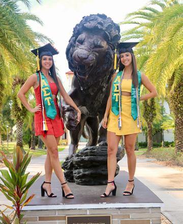 A photo of Jenna and Jessica Skrelunas, identical twins and Saint Leo alumni, smiling for a photo outside on University Campus next to the lion statue