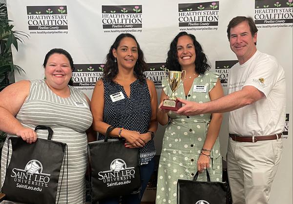 A photo of Mary Martinez-Drovie, Maria Dacosta, and Julia Sharp receiving their award and swag bags from Dr. Mark Gesner, vice president of Community Engagement and Communications at Saint Leo University, for placing first in the ‘A Solution That Matters’ category of the ‘What Matters on Mane Street’ social impact competition held by Saint Leo in 2022